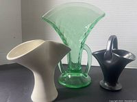 Three fan-shaped vases side by side against black background: green uranium glass in center, white ceramic on left, black glazed pottery on right.