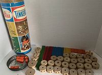 Full view of Tinkertoy wooden pieces next to original container showing about 80 pieces including various rods and spools, some damage visible.