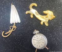 Front view of three vintage brooches: yellowy plastic dachshund, miniature metal alarm clock, and small umbrella brooch.