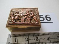 Top view of the brass trinket box with a ruler showing dimensions, featuring the cherub relief scene on the lid.