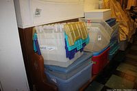 Stacked plastic storage tubs and containers including some with hinged lids, visible labels on clear tubs, mixed colors and sizes.