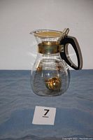 Clear glass coffee and tea maker carafe with black handle and gold-tone metal lid and collar. Gold starburst design visible on the glass. Tea ball placed inside the carafe.