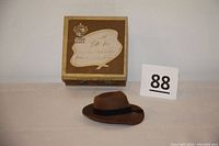 Photo showing small brown felt Adam Hat with black band and original square box with gift label on top.