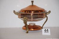 Side view of the assembled warming stand with copper lid on top showing the two handles, perforated copper band, glass bowl inside, and decorative legs.