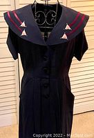 Full length front view of navy-blue sailor dress hung on dress form against white louvered doors. Shows dress shape, short sleeves, buttons, pockets, pleats, and distinctive collar with red stripes and white sailboats.