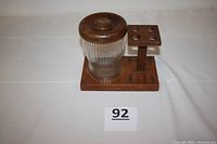 Ribbed glass humidor with wooden lid seated in wooden pipe stand, showing profile of the item with pipe slots and humidor.