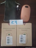 Two boxed rechargeable hand warmers with barcodes, one peach-colored hand warmer out of box, one black carry sleeve, one black USB charging cable.