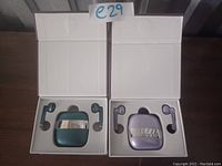 Opened packaging showing the purple and teal wireless earbuds with their respective charging cases and earbuds placed in foam cutouts.