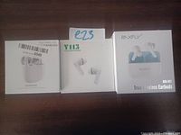 Photo showing three boxed sets of true wireless earbuds: one RAXFLY RAX-M01 and two unbranded sets, all in white cases with earbuds visible on packaging.