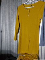 Photo showing a yellow ribbed long sleeved dress with round neckline and button details, hanging on a white hanger against a textured wall.