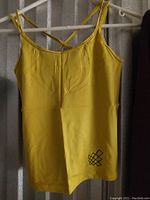 Yellow Jed North tank top hanging on a hanger against a plain background. Shows front view with crisscross straps visible at the top and small black geometric logo near bottom hem on left side.