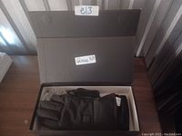 Photo showing a closed black rechargeable heated glove inside an open box placed on a wood surface. The glove size and lot number are indicated on white stickers.
