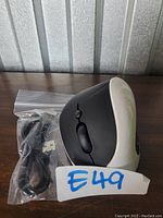 Black and white ergonomic right-hand mouse placed on a wooden surface with a sealed USB charging cable beside it.