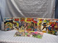 Front view of ten GI Joe comic books arranged standing and lying on surface, showing cover art of action scenes on each comic.