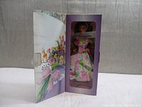 Barbie doll in pastel purple box with floral artwork opened showing the doll inside wearing floral dress and holding flowers.