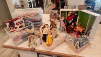 Overview of the lot showing multiple dolls, boxes, and containers with accessories and clothes displayed on a folding table in basement