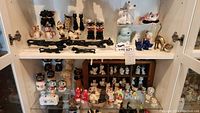 Front view of two shelves displaying multiple vintage cat-themed salt and pepper shakers and cat figurines arranged closely on white shelves in a cabinet.