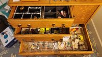 Top three drawers of oak jewelry chest showing various costume jewelry in divided trays including brooches, pins, bracelets, and rings.