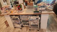 Crafting table top showing the white metal wire basket storage cabinets on wheels underneath, cluttered with various crafting supplies and materials.