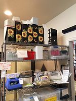 Three-tier metal wire shelving unit containing decorative magazine file holders, plastic storage bins, small glass jars, Cricut machines and manuals