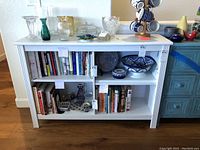 Full front view showing the white painted wood bookcase filled with books, several bowls, vases, and glassware on top and inside the shelves.