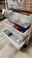 Three white wire mesh baskets stacked vertically, containing various plastic bins and containers inside with jewelry making supplies.
