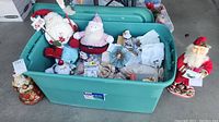 Green storage tote filled with assorted Santa-themed Christmas decor including plush dolls and other items, with a standing Santa figurine nearby.