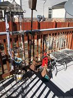 Wide photo showing patio railing with two vintage metal bistro chairs, red and green wooden birdhouses on the floor, metal plant stand with hanging pots, and ceramic pots arranged on the deck.