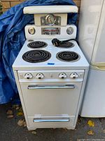 Front view of the white 1950s electric stove showing four coil burners and oven door with chrome handle, some rust spots and signs of use visible.