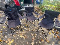Three black folding camping chairs positioned outdoors on ground covered with fallen leaves. Each chair has armrests with built-in drink holders and metal frame support.