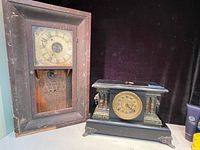 Photo shows two antique clocks; a tall wooden wall clock with a glass panel and a distressed face, and a black decorative mantel clock with a winding key on top.