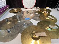 Photo showing 6 brass/bronze cymbals and a red-rimmed tambourine arranged on a table, exhibiting normal age wear.