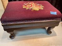 Side view of antique needlepoint footstool showing floral design on burgundy fabric and Queen Anne style wooden legs with curved feet.