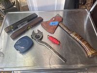 Photo showing antique tools including wooden level with two glass vials, red-handled axe with leather sheath marked #03, adjustable wrench, blue plastic tape measure, and red twine.
