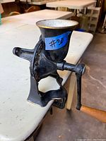 Side view of antique cast iron coffee grinder attached to table edge, showing black enamel body with white and gold-rimmed hopper and wooden handle crank