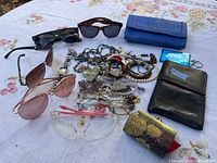 Lot containing assorted estate jewellery, sunglasses, wallets, coin purse, and a metallic watch on a tablecloth.