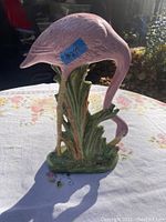 Figurine of a pink flamingo bending its neck downward on a green leafy base, placed on a floral tablecloth outdoors with sunlight creating shadows.