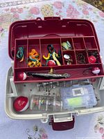 Open fishing tackle box showing assorted fishing lures, bobbers, hooks, and small plastic accessory box inside compartments
