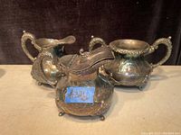 Set of three Victorian silver plated creamer, sugar bowl, and syrup pitcher on flat surface with dark background showing ornate embossed floral designs and ball feet