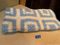 Photo showing folded handmade crochet blanket with blue and white square pattern. Dimensions about 37x37 inches.
