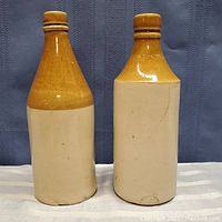 Two antique stoneware crock bottles side by side showing entire bodies with tan glazed top and off-white base, displayed on fabric