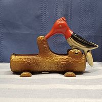 Side view of antique mechanical woodpecker toothpick server resting on brown textured log base with metal screw eye and detailed paint.