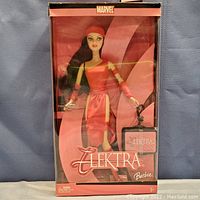 Front view of Barbie Elektra doll in original sealed box with pink and black themed packaging, showing doll in red outfit and Elektra branding.