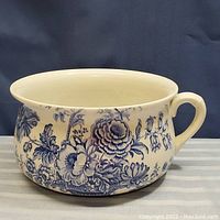 Front and side view of a porcelain chamber pot with blue floral transferware design on white background, showing one handle and wide open top, no lid present.