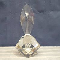 Clear crystal perfume bottle with geometric faceted body and stopper against a dark textured fabric background.