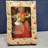 Boxed Christmas angel figurine shown through clear plastic window in original box decorated with Santa and star pattern.