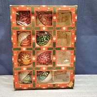 Front of original cardboard box showing 8 visible multicolored glass Christmas balls decorated with silver glitter snowflake patterns, and one empty compartment.
