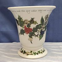 Front view of cream-colored ceramic vase decorated with holly and ivy botanical illustrations, showing red berries and green leaves, cursive text near rim