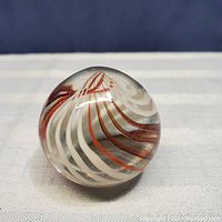 Spherical clear glass paperweight with internal swirl of red and white ribbons on white cloth background and blue backdrop.