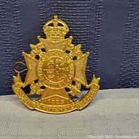 Front view of gold-tone metal Voltigeurs Canadian cap badge on textured background, showing crown, cross, and detailed crest with inscription.